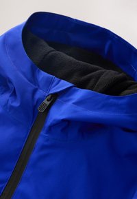 Veste bleue avec doublure polaire noire visible à l’intérieur du col et une fermeture éclair noire à l’avant, montrée sur un fond clair.