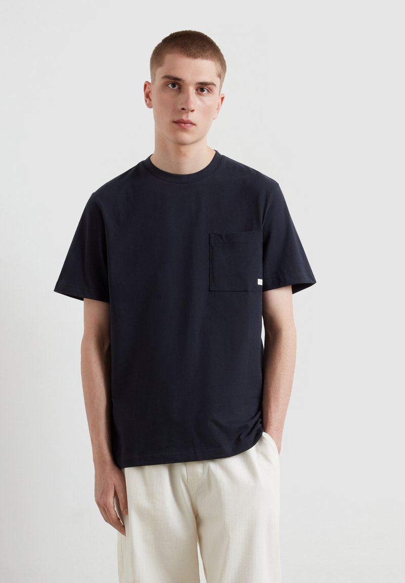 T-shirt in cotone blu navy con scollatura tonda e maniche corte, dotata di una tasca sul petto sinistro e una vestibilità rilassata. Design minimalista.