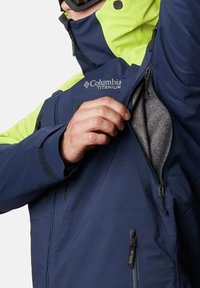 Giacca Columbia Titanium blu navy con accenti verde neon, dotata di una tasca sul petto con zip e una morbida fodera grigia. Cuciture dettagliate visibili.