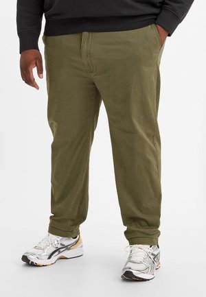 Personne portant un pantalon vert olive et des chaussures de sport blanches avec des touches noires et jaunes, debout avec une main dans la poche.
