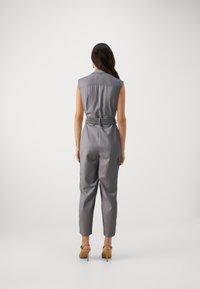 Ermeløs grå jumpsuit med belte i midjen, skreddersydd passform og subtil stripete tekstur. Sammen med tan høye hæler.