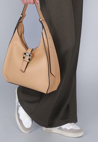 Osoba v černých kalhotách a bílých teniskách drží béžovou koženou kabelku s dekorativní sponou.