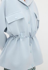 Chaqueta de forro polar azul claro con grandes bolsillos en los hombros, una cintura ajustable con cordón y botones a juego, que presenta un corte holgado y una textura suave.