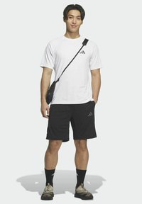 Maglietta sportiva bianca con un logo, pantaloncini neri e calzini neri. Una piccola borsa a tracolla nera è indossata sopra la spalla. Le scarpe sono beige e nere.