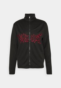 Giacca nera con zip realizzata in tessuto morbido, dotata di colletto alto e testo ricamato rosso sul davanti con un design frastagliato.