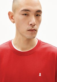 T-shirt en coton rouge avec un col rond, comportant un logo en ruban blanc sur la poitrine. Texture lisse avec des manches courtes.