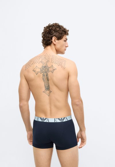 Tengerészkék boxeralsó világoskék derékpánttal, melyen logó látható. A modell hátán összetett mintákat ábrázoló tetoválás található.