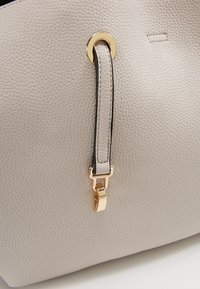 Lähikuva beigestä teksturoidusta nahkalaukusta, jossa on kultainen metallirengas ja hihnadetaali.