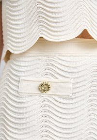 Ensemble en maille couleur crème avec texture ondulée, bords festonnés et un accent floral doré sur la ceinture. Tissu doux et côtelé partout.