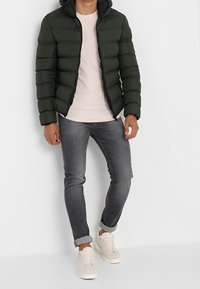 Grön puffjacka med huva, blixtlås och svarta accenter. Bärs över en ljusrosa skjorta och matchas med åtsittande gråjeans och vita sneakers.