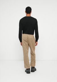 Uomo che indossa una camicia nera a maniche lunghe, pantaloni beige a fantasia e sneakers nere, in piedi di fronte a un muro bianco semplice.