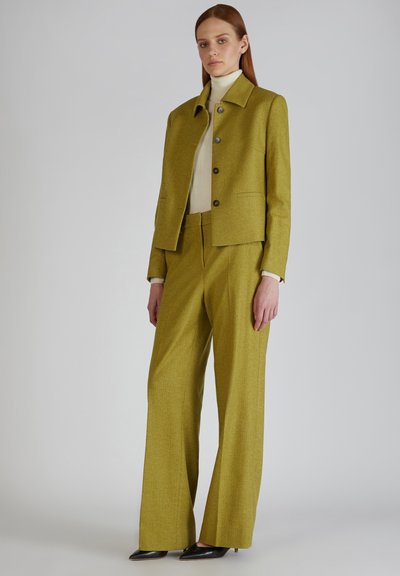 Groene gestructureerde suit met een boxy jasje, knoopsluiting en wijde broekspijpen, gecombineerd met een crèmekleurige coltrui en puntige zwarte hakken.