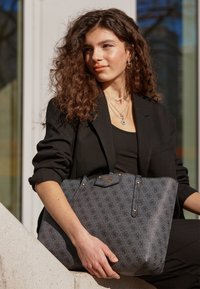 Stor sort totebag med tekstureret finish, der har et monogram-mønster, gyldne hardware-detaljer og et brandlogo på forsiden.