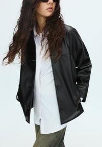 Schwarze Kunstlederjacke mit einem lockeren Schnitt, mit Knopfverschluss, großen Taschen und glatter Textur, die über einem hellen Hemd getragen wird.