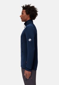 Marinblå fleecejacka med hög krage, dragkedjeförsedd ficka och logodetalj på vänster arm; slät textur, figursydd design.