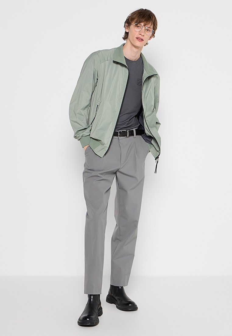 Junger Mann in einer hellgrünen Zip-Jacke, dunkelgrauem T-Shirt, grauen Hosen, schwarzem Gürtel, schwarzen Stiefeln und Brille, der vor einem weißen Hintergrund steht.