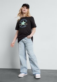 Camiseta negra con un gráfico de estrella verde y texto; vaqueros anchos de piernas deshilachadas en azul claro; zapatillas blancas con acentos rojos y gorra negra.