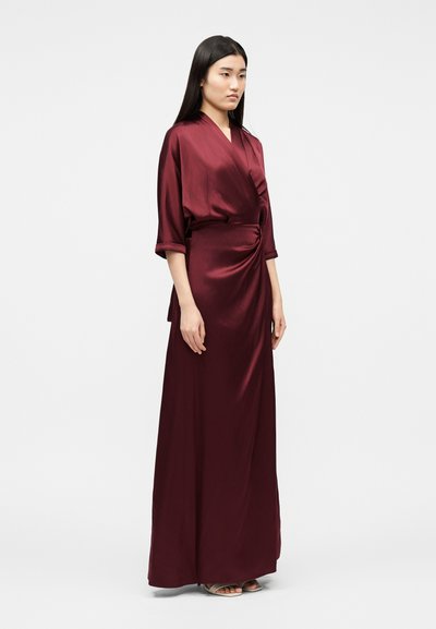 Γυναίκα που στέκεται με τα χέρια στα πλευρά, φοράει μακρύ φόρεμα σατέν σε βαθύ μπορντό χρώμα, με μανίκια τριών τετάρτων και τύλιγμα στη μέση.