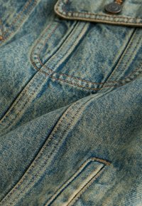 Közeli kép világoskék farmer anyagról, kettős narancssárga varrással és egy fémből készült gombbal a zsebfedőn.