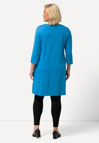 Blaue langärmlige Tunika mit geradem Schnitt, kombiniert mit schwarzen Leggings und Pumps, von hinten betrachtet. Glatte Stoffoberfläche.