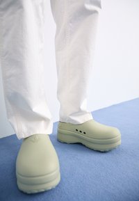 Gröna gummiklumpor med en jämn yta, rundad form och tre ventilationshål; kombinerade med vita byxor på en blå yta.