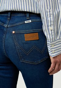 Jeans in denim blu scuro con un'etichetta in pelle che presenta il logo "Wrangler" sulla tasca posteriore. La manica di una maglietta a righe è visibile accanto ai jeans.