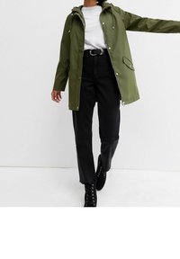 Oliwkowa wodoodporna kurtka z kapturem, zapinana na zamek i zatrzaski, zestawiona z czarnymi dżinsami z wysokim stanem oraz czarnymi sznurowanymi botkami.