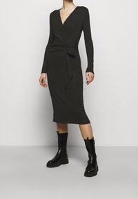 Abito avvolgente nero in maglia con maniche lunghe, scollatura a V profonda e dettaglio con nodo laterale, abbinato a stivali neri con tacco alto fino al ginocchio.