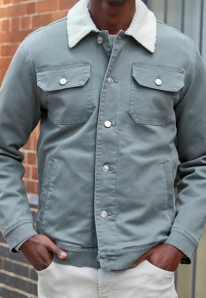 Homme portant une veste grise boutonnée avec un col en polaire blanche, les mains dans les poches, debout devant un mur en briques.