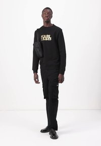 Sudadera negra con el texto dorado "KARL LAGERFELD", combinada con pantalones cargo negros y un pequeño bolso cruzado negro. El modelo se encuentra de pie frente a un fondo blanco.