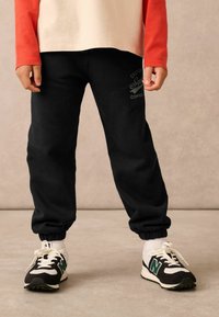Schwarze Baumwoll-Jogginghosen mit elastischen Bündchen und einem Logodetail an den Oberschenkeln, kombiniert mit schwarz-weißen Turnschuhen mit grünen Akzenten.