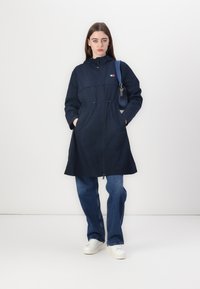 Cappotto impermeabile blu navy con vita regolabile, tasche frontali e cappuccio; abbinato a jeans azzurri e sneakers bianche.