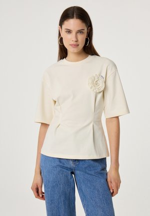 Woman wearing a cream short-sleeve top with a textured flower detail on the chest, paired with blue denim jeans.