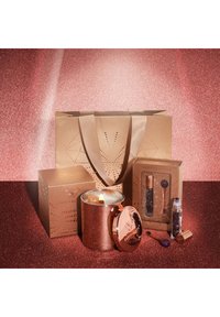 Set regalo in oro rosato che include una candela in un portacandela metallico, due articoli in scatola e una borsa con dettagli perforati, il tutto su uno sfondo glitterato.