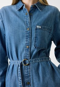 Abito camicia di jeans azzurro chiaro con colletto classico, chiusura frontale con bottoni, taschino sul petto e cintura in tessuto con fibbia rotonda dorata.