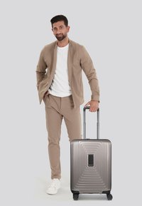 Valigia rigida di colore grigio metallizzato con una superficie pattern e un design elegante, posizionata su quattro ruote, accanto a una persona in un outfit beige.