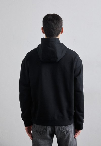 Black hooded sweatshirt with a soft texture, relaxed fit, and ribbed cuffs. Worn with gray jeans, showing stitching details.