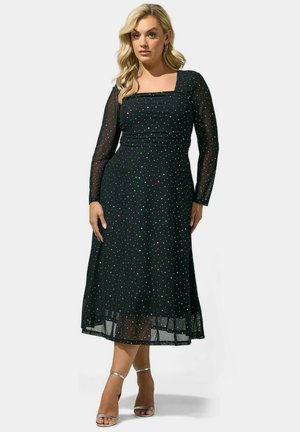 Femme blonde portant une robe midi noire à manches longues transparentes avec des pois multicolores, associée à des talons ouverts argentés.