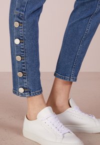 Jeans de mezclilla azul con cinco botones plateados en el dobladillo exterior, combinados con zapatillas blancas. Diseño de pierna recta con detalle de costura visible.