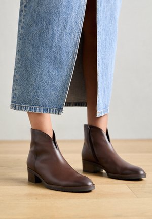 Persona con falda de mezclilla azul claro de longitud media y botas de cuero marrón hasta el tobillo, de pie sobre un suelo de madera.