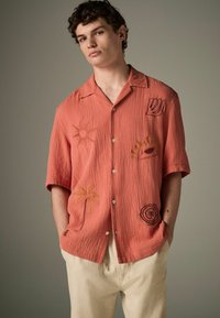 Kortærmet t-shirt i koralfarvet bomuld med tekstureret stof; har broderede detaljer som sol, palme, øje og andre dekorative elementer. Design med knaplukning foran.