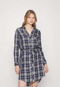 Abito camicia a quadri navy con maniche lunghe, chiusura con bottoni, tasca sul petto e cintura da annodare. Realizzato in un tessuto morbido e leggero.