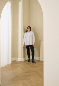 Femme aux longs cheveux bruns portant un chemisier blanc, un pantalon noir et des bottes noires, debout dans une pièce beige avec une porte voûtée et un sol en parquet en bois.