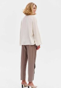 Lockere cremefarbene Bluse, kombiniert mit grauen Hosen mit seitlichen Schlitzen. Das Model trägt hellfarbige Absatzmules. Glatter Stoff mit angenehmer Textur.