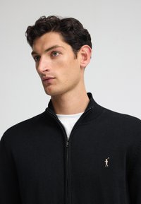 Svart zip-up sweatshirt med rilad krage, prydd med en liten, diskret broderad logotyp på bröstet, tillverkad av strukturerat stickat material.