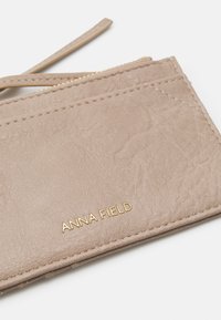 Beige kortholder laget av teksturert syntetisk lær, med en gull "ANNA FIELD"-logo, glidelåslomme og sømkanter.
