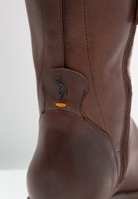 Botte en cuir marron avec une fermeture éclair latérale, une surface texturée, et un accent de design noir avec un logo d'oiseau près du haut.