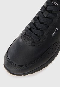 Black HUGO sneaker with textured toe cap, black laces, and branded side panel detail on white background.