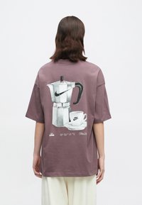 Oseba s približno dolžino do ramen in rjavimi lasmi nosi preveliko mauve majico z grafiko moka lonca in skodelice kave, ki ima logotipe Nike.