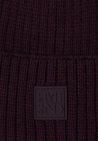 Tissu en tricot côtelé violet foncé avec une étiquette rectangulaire cousue, présentant des lettres stylisées et un design en relief.